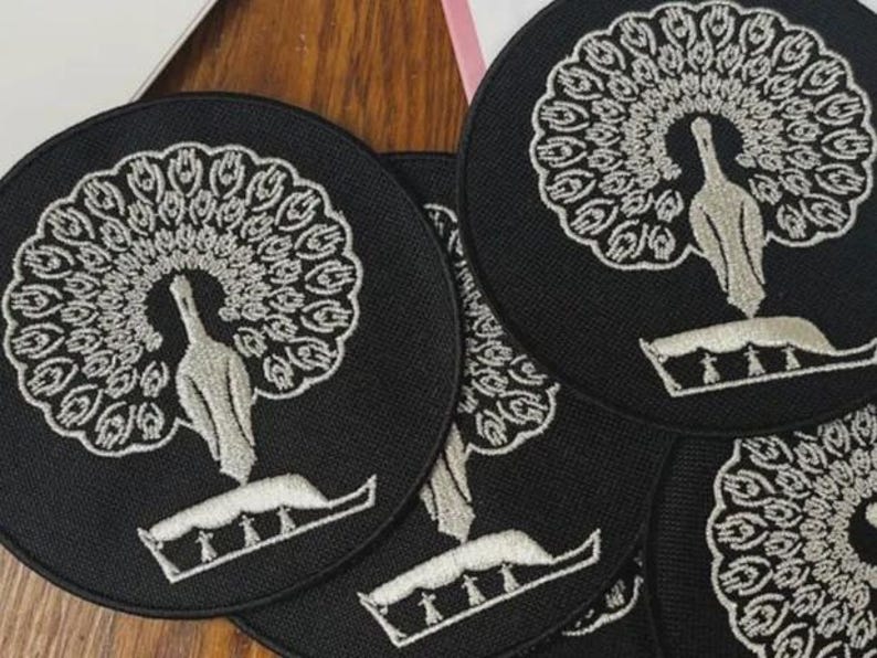 Pu&ograve; includere: Una collezione di toppe circolari in tessuto nero con un ricamo raffigurante un pavone color argento. Ogni toppa presenta un pavone stilizzato con le piume della coda spiegate a ventaglio circolare sopra un elemento di base decorativo.