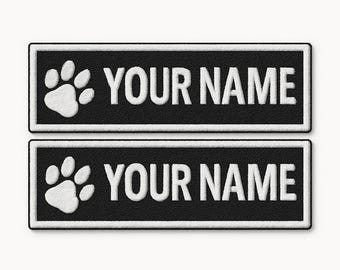 Può includere: Due toppe rettangolari nere con bordi ricamati bianchi. Ogni toppa presenta un'icona di impronta di zampa bianca sul lato sinistro e il testo bianco "YOUR NAME" sul lato destro, in un carattere sans-serif chiaro e in grassetto.