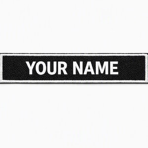 Pode incluir: Um emblema retangular bordado em preto com uma borda branca. O texto "YOUR NAME" está costurado em letras maiúsculas brancas no centro do emblema. O design apresenta um visual limpo e de alto contraste com um acabamento de tecido texturizado.