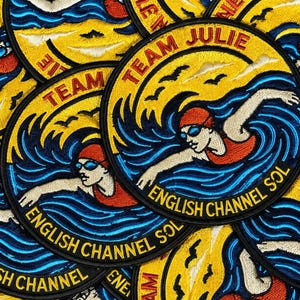 Pu&ograve; includere: Una toppa ricamata circolare raffigurante una persona che nuota con cuffia e costume rossi tra onde blu. Lo sfondo giallo mostra uccelli neri e il testo TEAM JULIE in alto e ENGLISH CHANNEL SOL in basso in nero.