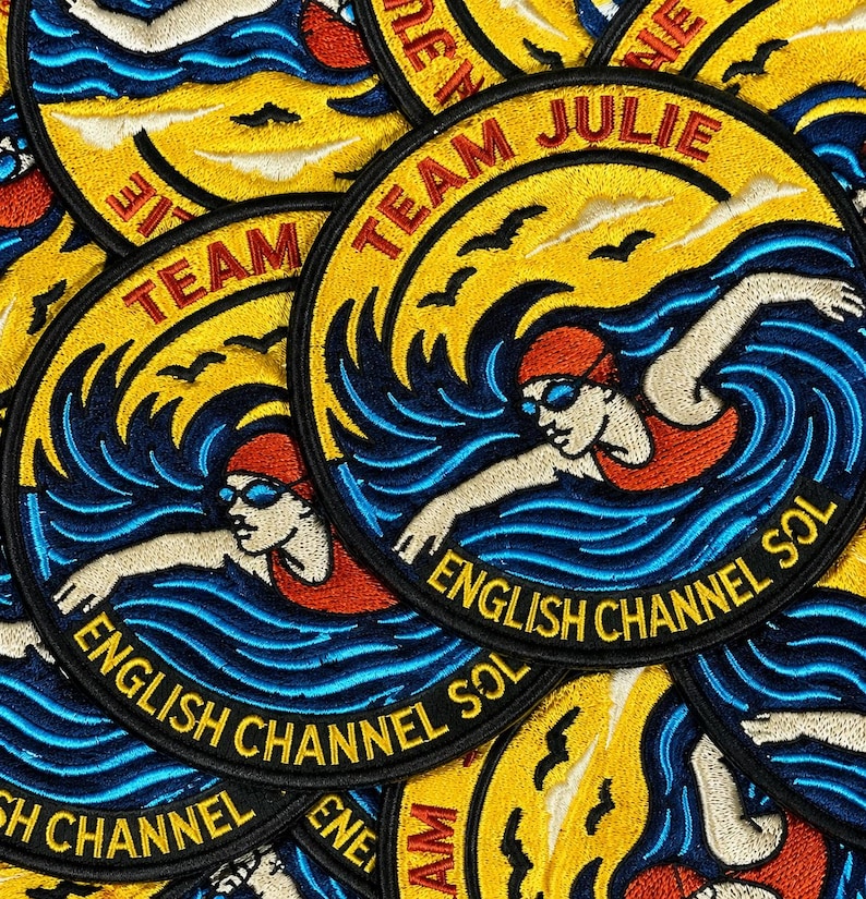 Pu&ograve; includere: Una toppa ricamata circolare raffigurante una persona che nuota con cuffia e costume rossi tra onde blu. Lo sfondo giallo mostra uccelli neri e il testo TEAM JULIE in alto e ENGLISH CHANNEL SOL in basso in nero.