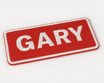 Può includere: Una toppa rettangolare ricamata con sfondo rosso e bordo bianco. Il nome GARY è cucito in lettere maiuscole bianche e in grassetto al centro della toppa. La trama del ricamo è visibile su tutta la superficie dell'oggetto.