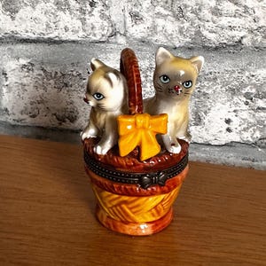 Op de afbeelding: Een keramische sieradendoos in de vorm van een mand, met twee kleine kattenbeeldjes. De mand is bruin en geel met een oranje strik. De katten zijn bruin en wit met blauwe ogen.