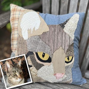 May include: A decorative square pillow featuring a patchwork cat face design. The cat's face is composed of various textured fabrics in shades of gray, brown, and beige, with yellow eyes and a pink nose. The pillow has a blue denim background and a plaid corner.
