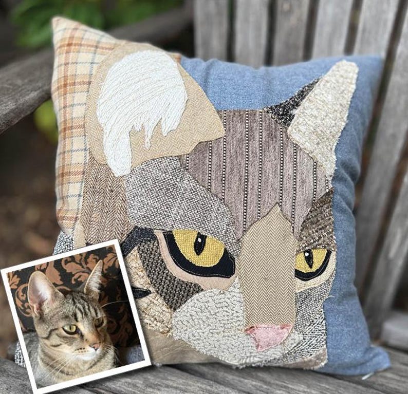 May include: A decorative square pillow featuring a patchwork cat face design. The cat's face is composed of various textured fabrics in shades of gray, brown, and beige, with yellow eyes and a pink nose. The pillow has a blue denim background and a plaid corner.