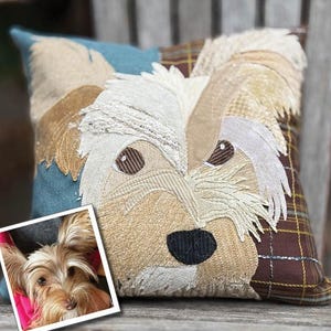 May include: Square decorative pillow featuring a dog's face. The pillow has a patchwork design with various textures and colors, including beige, brown, blue, and plaid. The dog's face is detailed with embroidered features and a black nose. A small photo of the dog is in the corner.