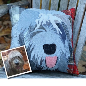 May include: A decorative square pillow featuring a dog face design. The pillow has a light blue and gray color scheme with a black nose and pink tongue. A framed photo of a dog is in the lower left corner. The pillow is placed on a wooden bench.