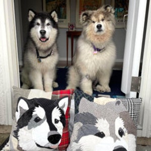 May include: Two fluffy Alaskan Malamute dogs sit inside a doorway, above two decorative pillows with dog faces. The pillows are patchwork with black, white, and gray fabrics. The dogs wear collars. Artwork and a small table are visible in the background.