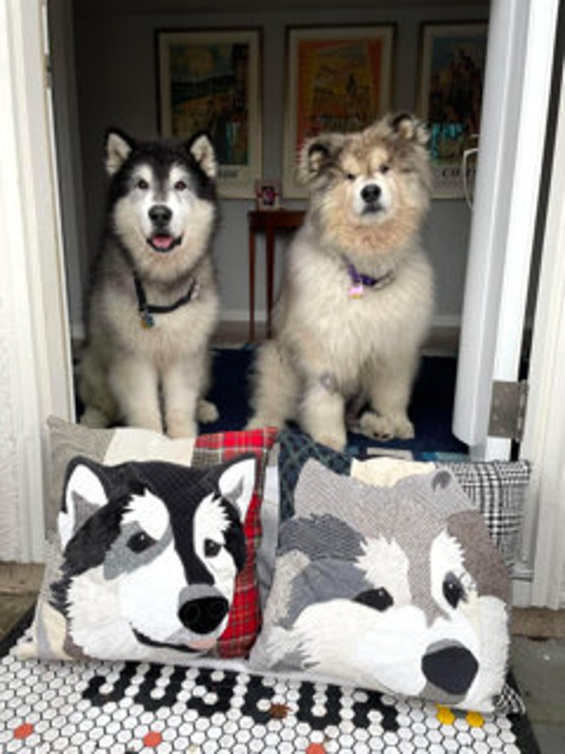 May include: Two fluffy Alaskan Malamute dogs sit inside a doorway, above two decorative pillows with dog faces. The pillows are patchwork with black, white, and gray fabrics. The dogs wear collars. Artwork and a small table are visible in the background.