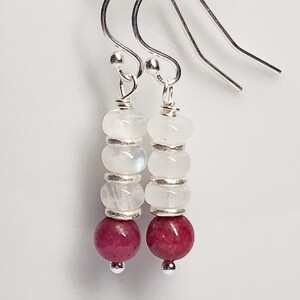 May include: A pair of silver dangle earrings with white moonstone beads and a single red ruby bead at the bottom.