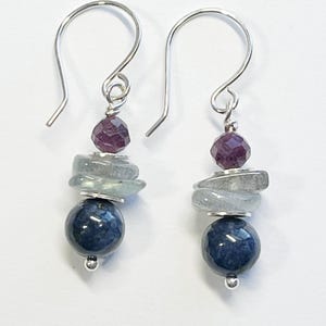May include: A pair of dangle earrings with silver hooks. Each earring features a dark blue sphere, a stack of light blue-green stones, and a faceted purple bead. The earrings are designed with a minimalist style.