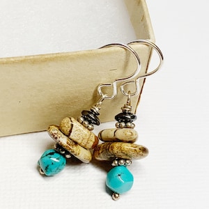 May include: Handmade earrings featuring turquoise and stacked stone beads. The earrings have silver-tone hooks and accents. The stones are a mix of tan and brown, with a pop of bright turquoise. The earrings are presented in a neutral-toned box.