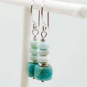 May include: A pair of dangle earrings with a silver hook. Each earring features a stack of turquoise, light blue, and white beads. The bottom bead is a larger, square-cut turquoise stone. The earrings are displayed on a white stand.
