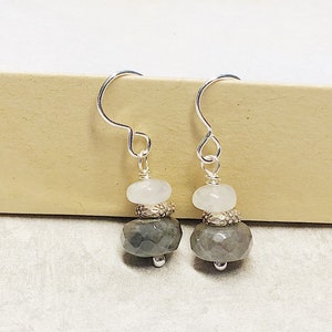 May include: Silver dangle earrings with a gray and white gemstone design. The earrings feature a silver hook and a gray gemstone bead with a white gemstone bead above it.