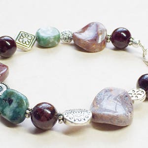 May include: A beaded bracelet featuring a variety of natural stones and silver-toned accents. The bracelet includes polished maroon, green, and patterned brown stones, interspersed with silver beads and decorative metal pieces. The bracelet is secured with a silver clasp.