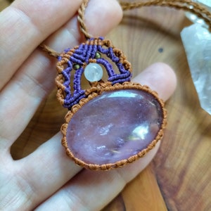 May include: A purple and brown macrame necklace with a large oval-shaped rose quartz pendant. The pendant is surrounded by a brown macrame design and a smaller white quartz bead.