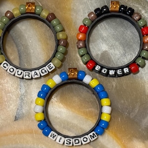 May include: Three circular bracelets featuring black bands adorned with colorful plastic beads. One bracelet has green beads and the word COURAGE, one has red, orange, and green beads with the word POWER, and one has blue, yellow, and white beads with the word WISDOM.
