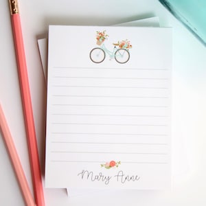 May include: A white notepad with lined paper and a floral design featuring a bicycle with a basket of flowers. The notepad has the name "Mary Anne" printed at the bottom.