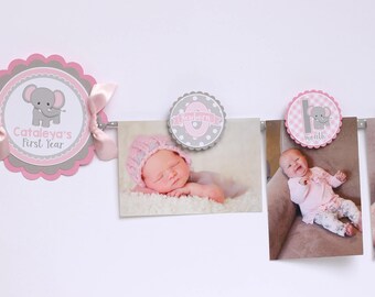 May include: A baby's first year photo banner with pink and gray accents. The banner includes photos of a baby at different ages, with decorative circles featuring an elephant design and the baby's name. The banner is strung on a gray ribbon.