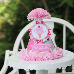 May include: A pink and white ice cream cone party hat with the word "one" and the name "Maddie" on it. The hat is decorated with pink, white, and blue paper.
