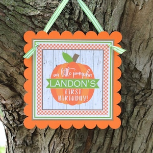 May include: Orange and green pumpkin-themed sign with scalloped edges. The sign reads "our little pumpkin LANDON'S first birthday!"