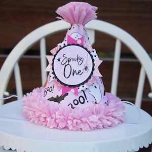 May include: A pink and white Halloween-themed party hat with the words "Spooky One" and "Boo!" printed on it. The hat features a spider web design, a spider, and ghost illustrations. It is decorated with pink tissue paper.