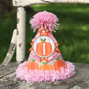 May include: A pink and orange pumpkin-themed party hat with a pink pom-pom on top. The hat has a white circle with a number "1" and a pumpkin on it. The hat is sitting on a wooden chair.