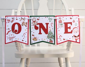 Könnte beinhalten: Ein weißer Hochstuhl mit einem rot-grün-weißen Weihnachtsbanner, auf dem in roten Buchstaben "ONE" steht. Das Banner zeigt verschiedene Weihnachtsmotive, darunter einen Schneemann, ein Rentier, einen Lebkuchenmann, Zuckerstangen und einen Weihnachtsmann.
