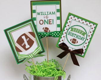 May include: Three green and brown cardstock signs with a football theme. The signs say "William is One!" "Happy Birthday" and "1" with a football graphic. The signs are on wooden skewers and are in a silver bucket with green shredded paper.