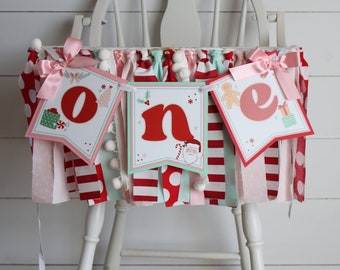 Könnte beinhalten: Ein rot-weiß-grünes Weihnachtsbanner mit der Aufschrift "eins" mit Motiven von Weihnachtsmann, Lebkuchenmann und Geschenken. Das Banner hängt an einem weißen Hochstuhl.