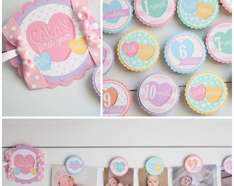 May include: A pink and purple baby milestone banner with a heart design. The banner includes a title card that says "Cara's First Year" and individual cards for each month of the year. The banner is decorated with white polka dots and pink bows.