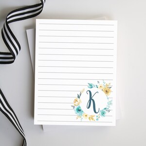 May include: A white lined notepad with a floral wreath design featuring a blue letter K in the center. The wreath is made up of yellow and blue flowers and green leaves.