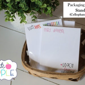 May include: Three white note cards with colorful text, including "a note from MS. ETTELI", "MRS. dangel", and "teach love inside". The cards are in a brown wicker basket with a cellophane sleeve. The text "pink APPLE" is visible in the lower left corner of the image.
