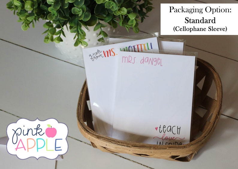 May include: Three white note cards with colorful text, including "a note from MS. ETTELI", "MRS. dangel", and "teach love inside". The cards are in a brown wicker basket with a cellophane sleeve. The text "pink APPLE" is visible in the lower left corner of the image.