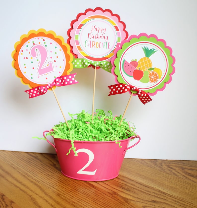 May include: A pink metal bucket with the number "2" on the front, filled with green shredded paper. Three colorful paper circles with scalloped edges are on sticks in the bucket. The circles say "Happy Birthday Caroline", "2" with colorful polka dots, and a fruit design.