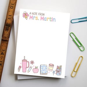 May include: White notepad with the text "A Note From Mrs. Martin" in pastel colors. The notepad has a decorative border with school-themed illustrations, including a pencil, apple, and coffee cup. A wooden ruler and colorful paper clips are in the background.