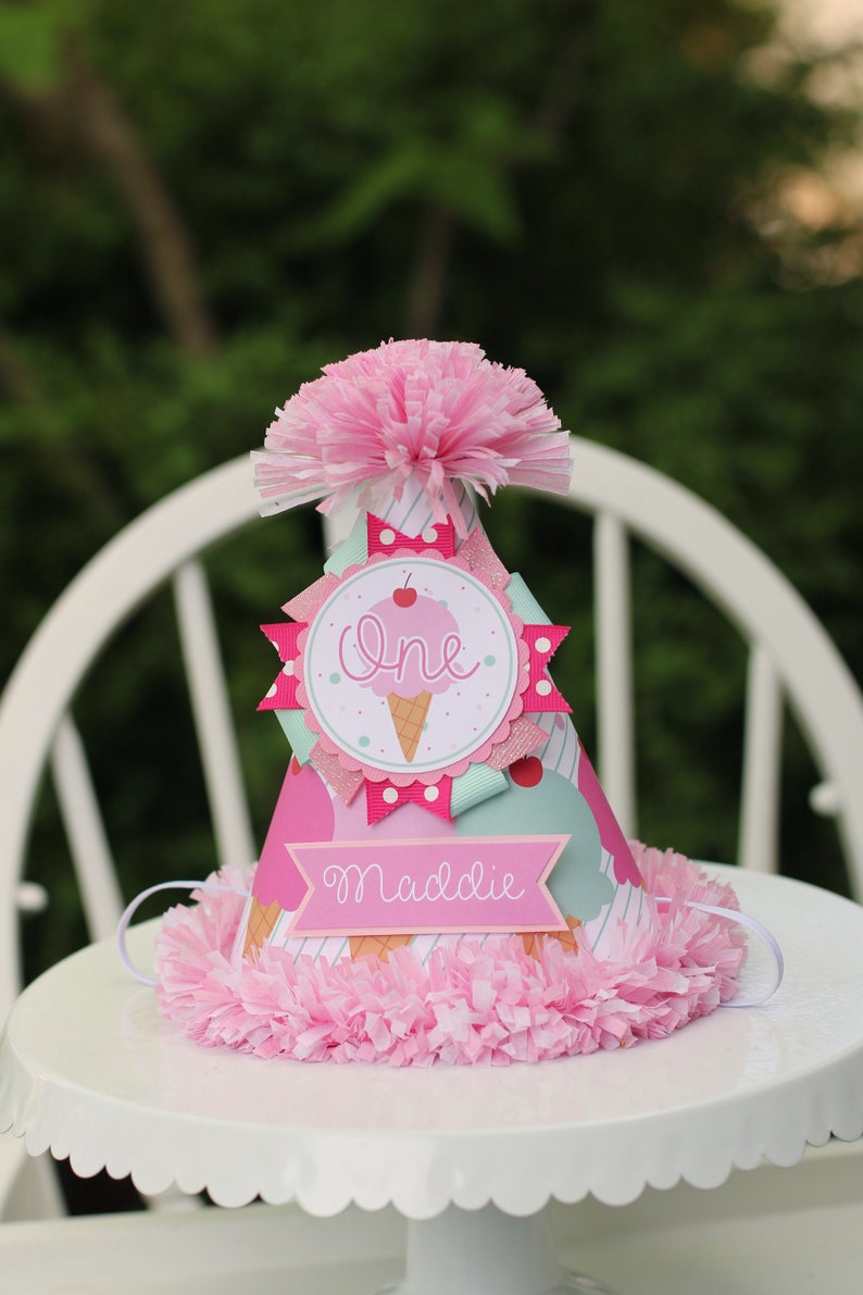 May include: Pink and white birthday party hat with an ice cream cone design. The hat says "One" and "Maddie" on it.