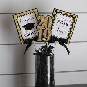 Puede incluir: Un centro de mesa de graduaci&oacute;n con decoraciones negras y doradas. El centro de mesa incluye un jarr&oacute;n lleno de papel negro triturado, tres letreros con el texto "Congrats Grad", "2019" y "Class of 2019 Angie", y una cinta negra y dorada.