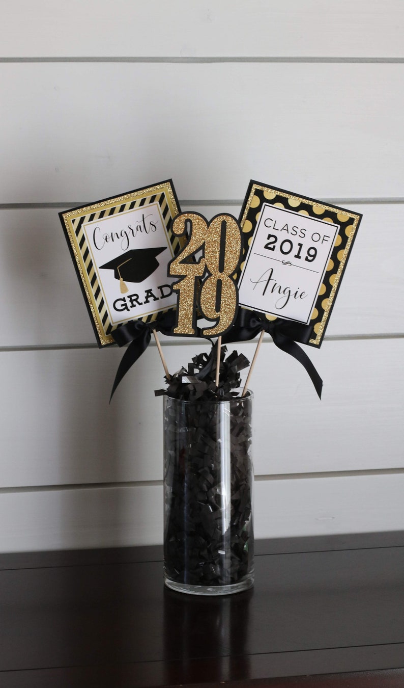 Puede incluir: Un centro de mesa de graduaci&oacute;n con decoraciones negras y doradas. El centro de mesa incluye un jarr&oacute;n lleno de papel negro triturado, tres letreros con el texto "Congrats Grad", "2019" y "Class of 2019 Angie", y una cinta negra y dorada.