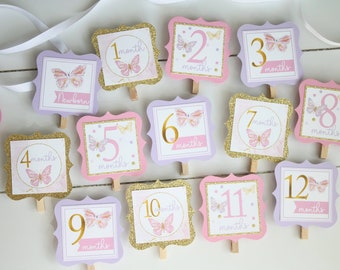 May include: Twelve pink and purple square cards with gold glitter and butterfly designs. Each card has a number from 1 to 12 with the word "months" written below. The cards are attached to wooden clothespins.