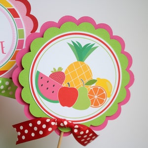 May include: A green and pink scalloped circle with a white circle inside. The white circle has a colorful illustration of a pineapple, watermelon, strawberry, apple, pear, and orange. The text "Happy birthday Caroline" is on the other circle.