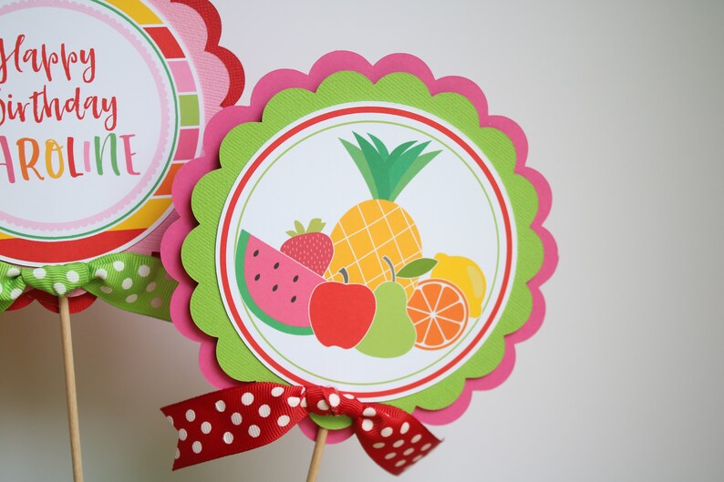 May include: A green and pink scalloped circle with a white circle inside. The white circle has a colorful illustration of a pineapple, watermelon, strawberry, apple, pear, and orange. The text "Happy birthday Caroline" is on the other circle.
