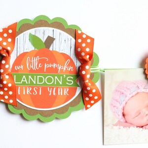 Puede incluir: Una pancarta en forma de calabaza verde, marr&oacute;n y naranja con el texto "our little pumpkin Landon's first year" y un lazo blanco y naranja con lunares. Una foto de un beb&eacute; con un gorro rosa est&aacute; unida a la pancarta.