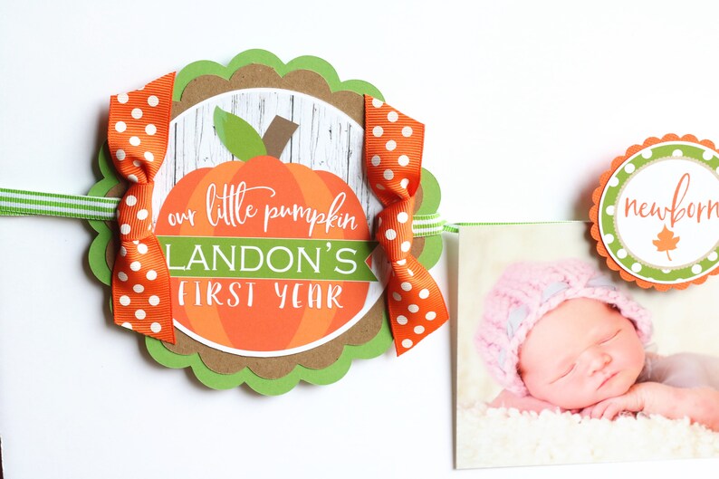 Puede incluir: Una pancarta en forma de calabaza verde, marr&oacute;n y naranja con el texto "our little pumpkin Landon's first year" y un lazo blanco y naranja con lunares. Una foto de un beb&eacute; con un gorro rosa est&aacute; unida a la pancarta.