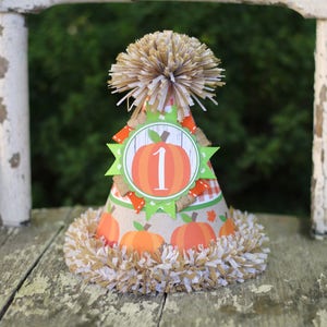 Puede incluir: Un gorro de fiesta de papel marr&oacute;n y blanco con una calabaza y el n&uacute;mero "1" en &eacute;l. El gorro tiene un pomp&oacute;n marr&oacute;n y blanco en la parte superior y una franja marr&oacute;n y blanca alrededor de la parte inferior.