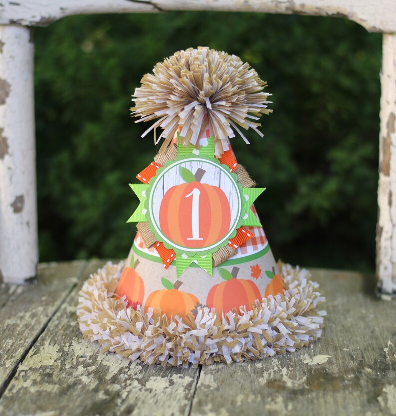 Puede incluir: Un gorro de fiesta de papel marr&oacute;n y blanco con una calabaza y el n&uacute;mero "1" en &eacute;l. El gorro tiene un pomp&oacute;n marr&oacute;n y blanco en la parte superior y una franja marr&oacute;n y blanca alrededor de la parte inferior.