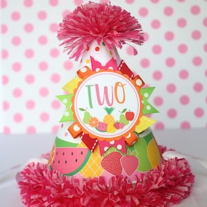 May include: A pink and white party hat with a fruit design and the word "TWO" in a circle. The hat has a pink pom-pom on top and a pink and white fringe around the bottom.
