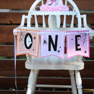 May include: A white wooden high chair decorated for a Halloween-themed first birthday. The chair has a banner that spells "ONE" with spooky graphics and a matching party hat. The banner has a ghost, spider, and spiderweb.