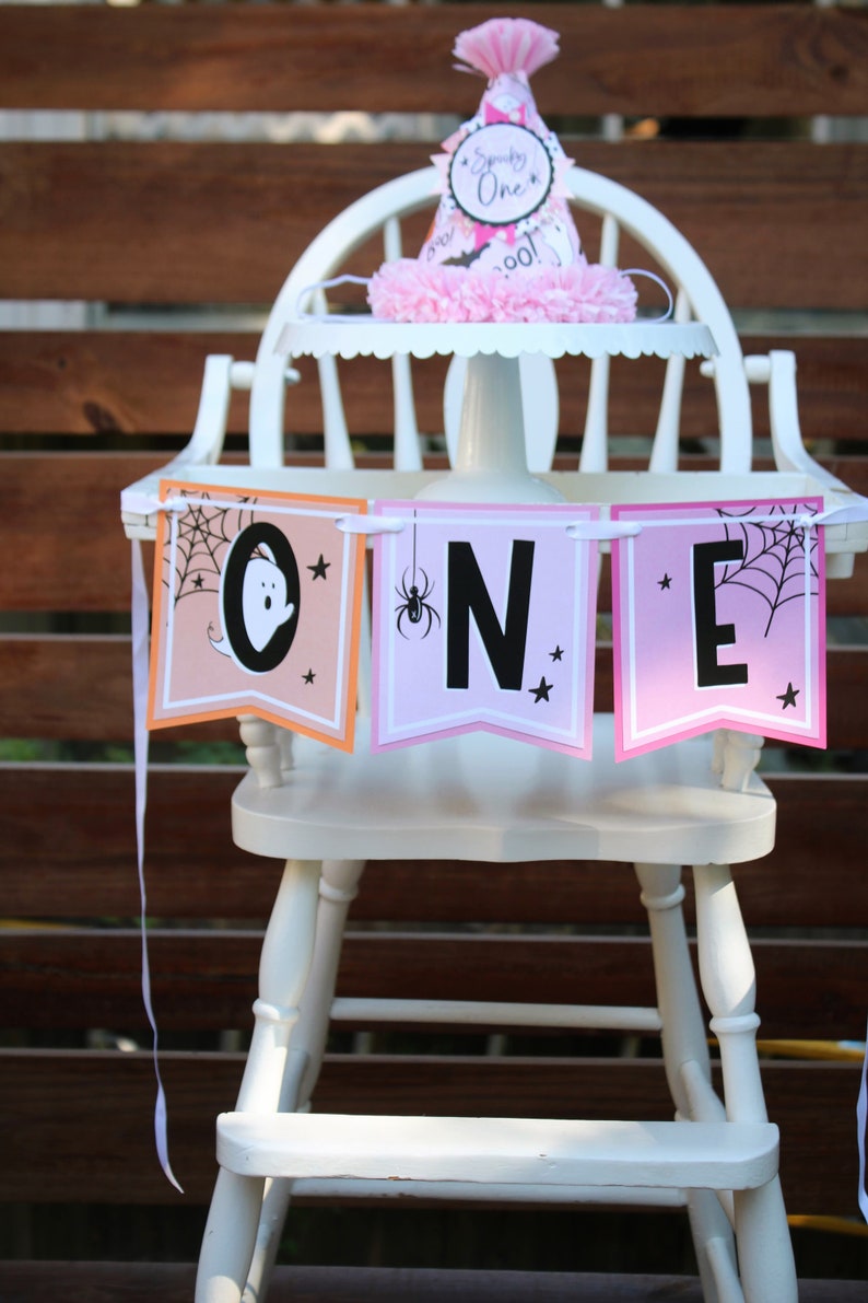 May include: A white wooden high chair decorated for a Halloween-themed first birthday. The chair has a banner that spells "ONE" with spooky graphics and a matching party hat. The banner has a ghost, spider, and spiderweb.
