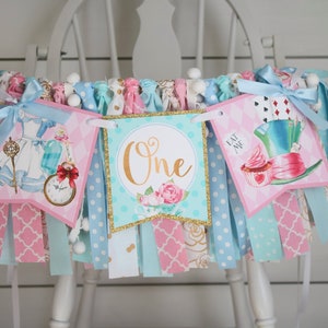 May include: A pink and blue Alice in Wonderland themed birthday banner with the word "One" in gold glitter. The banner features images of Alice, the Mad Hatter, and a cupcake.